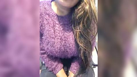 Media: A video of a woman with long, wavy brown hair, wearing a fuzzy, purple sweater, seated indoors, with a blurred, purple background.