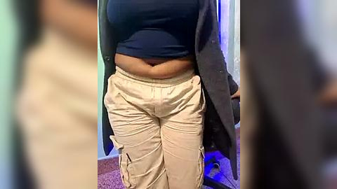 Media: Video of a person with dark skin, wearing a blue crop top, loose beige cargo pants, and a grey cardigan, standing in a dimly lit room with a carpeted floor.