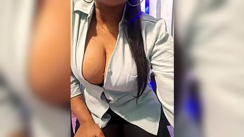 Media: Video of a woman with medium skin tone, long black hair, wearing a partially unbuttoned white blouse revealing cleavage, sitting indoors with blurred background.