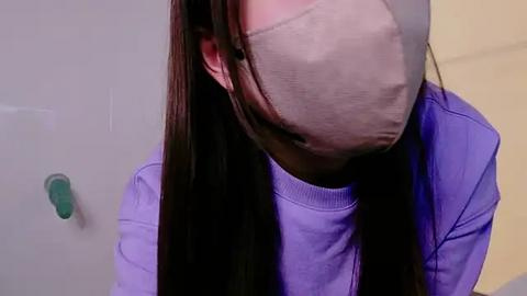 Media: Video of a person with long, straight black hair wearing a light gray face mask and a purple shirt, standing against a pale yellow and white wall, focusing on their head and shoulders.