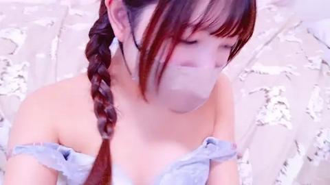 Media: Video of an Asian woman with light skin, wearing a light blue off-shoulder top, with a braided red-brown ponytail. She has a white face mask on and is sitting against a white, textured background.