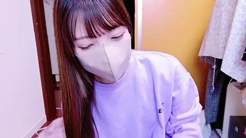 Media: A video of an Asian woman with long brown hair and pale skin, wearing a white t-shirt and beige face mask, kneeling on a tiled floor in a dimly lit room with a wooden door and hanging clothes.