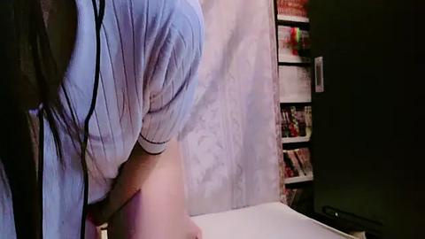 Media: A video of a person with light blue hair, wearing a white shirt, bending over a white table, in a dimly lit room with bookshelves in the background.