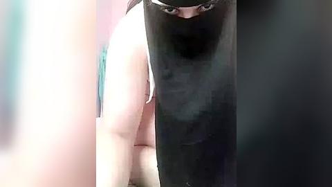 Media: Video of a person with a black hood obscuring their face, standing in a dimly lit room with blurred, soft-colored walls.