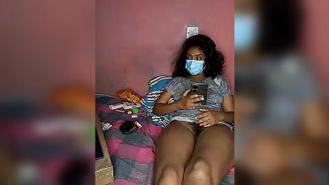 Media: Video of a young Black woman with dark curly hair and light brown skin, wearing a blue face mask, grey T-shirt, and blue shorts, sitting on a colorful striped bed, holding a smartphone, in a dimly lit room with pink walls.