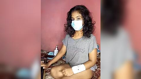 Media: Video of a young woman with curly black hair, wearing a face mask, grey t-shirt, white bandage on her right wrist, seated on a patterned bedspread, in a pink-walled room.