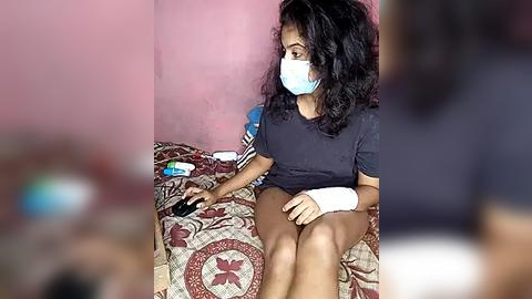 Media: Video of a young woman with dark curly hair, wearing a blue face mask, black T-shirt, and white cast on her right arm, sitting on a bed with a red and white patterned blanket. The background is a pink wall.