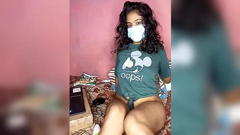 Media: Video of a young, dark-skinned woman with curly hair and a mask, wearing a \"Pops!\" t-shirt and no pants, sitting on a bed in a pink room, with a blurred background.