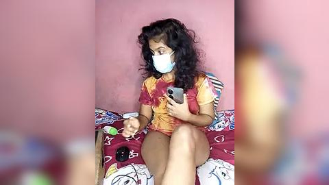 Media: Video of a young woman with dark curly hair, wearing a red tie-dye shirt, face mask, and holding a smartphone, sitting on a bed with a colorful, cartoon-patterned duvet.