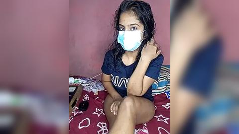 Media: Video of a young South Asian woman with medium skin tone, curly dark hair, wearing a blue face mask and dark T-shirt, sitting on a bed with a red and white star-patterned blanket.