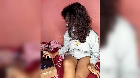 Media: Video of a young girl with curly dark hair, wearing a light grey sweatshirt and pink shorts, seated on a red cushion, surrounded by messy clothes on a pink wall.