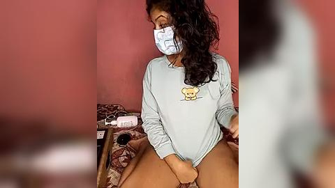 Media: Video of a South Asian woman with medium-dark skin, wearing a light blue sweatshirt, white mask, and tan shorts, sitting on a bed with a red wall and patterned bedding, holding her crotch.