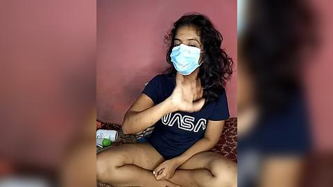 Media: Video of a woman with medium skin tone, curly black hair, wearing a blue face mask, navy blue t-shirt, sitting cross-legged on a bed with a red blanket, in a pink room, touching her cheek.