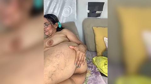 Media: Video of a plus-size woman with glasses, dark hair, and green scrunchie, lying on a bed, nude, with her hand on her large, round buttocks. Background shows a gray couch, yellow pillow, and white wall with a vent.