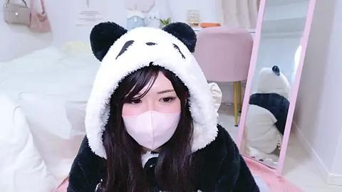 Media: Video of an Asian woman with long black hair in a panda costume, wearing a face mask, sitting in a cozy, pastel-colored bedroom with a pink chair and white floor.