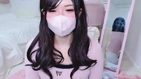 Media: Video of an Asian woman with long, wavy black hair, wearing a white face mask and a pink knit top. She sits on a bed with a pink blanket in a softly lit, minimalist bedroom.