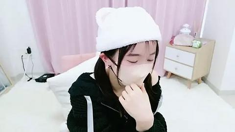 Media: Video of an East Asian woman with long black hair, wearing a white knit beanie, face mask, and black hoodie, sitting on a white bed in a minimalist room with pink curtains, wooden nightstand, and a green plant.