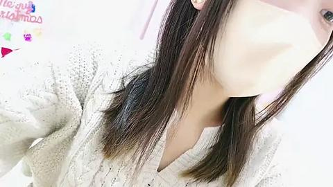 Media: Video of a young Asian woman with long, straight dark brown hair, wearing a beige knitted sweater and a face mask. Background shows a blurred, white room.