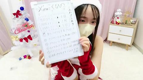 Media: Video of an Asian woman in a Santa costume, holding a Christmas list, in a festive room with a decorated tree, gifts, and a wooden nightstand.