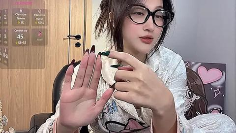 Media: Video of an Asian woman with fair skin, dark hair, and black-rimmed glasses, wearing a white, patterned robe, applying green nail polish on her fingers in a cozy, well-lit room.