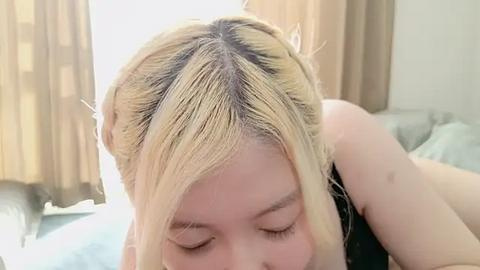 Media: Video of a young Asian woman with platinum blonde hair, eyes closed, leaning over a bed, wearing a black top. Background includes beige curtains and a light-colored bedspread.