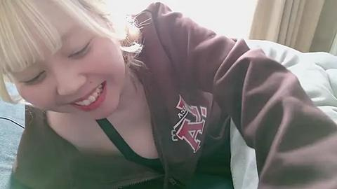 Media: A video of a smiling Asian woman with blonde hair and light skin, wearing a brown jacket with a large \"TSA\" logo, lying on a bed with white sheets and beige curtains in the background.