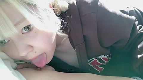 Media: Video of a young girl with blonde hair, blue eyes, and light skin, sticking out her tongue, wearing a dark hoodie with a red and white logo, lying on a bed with white sheets.