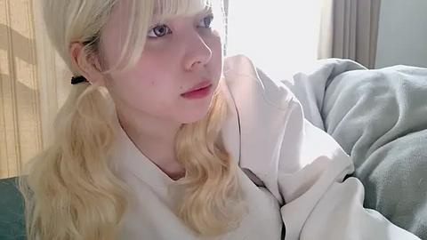 Media: Video of a young Caucasian woman with fair skin, wavy blonde hair in pigtails, wearing a white robe, sitting on a couch, looking contemplative, in a softly lit room with beige curtains.