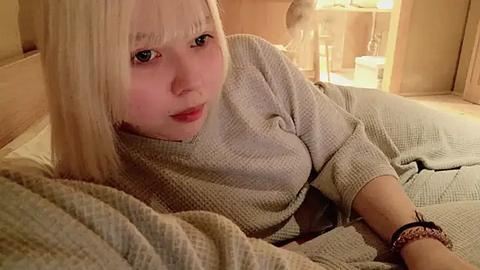 Media: Video of a fair-skinned, blonde woman in a beige knit sweater, lying on a bed in a dimly lit, cozy bedroom with wooden furniture and soft lighting.