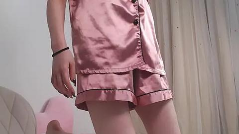 Media: Video of a person wearing a pink satin pajama set with a buttoned front and pleated shorts. The background features a beige curtain and a blurred, pink object on the left.