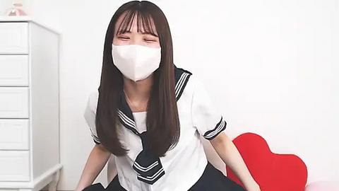 Media: Video of an Asian woman with long black hair, wearing a white mask, school uniform, and black skirt, seated on a red heart-shaped cushion. Background includes white drawers and plain white wall.