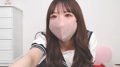 Media: Video of an East Asian woman with long brown hair and bangs, wearing a white sailor school uniform and pink surgical mask, reaching out with both hands. Background shows a white dresser and a pink balloon.