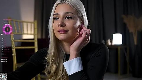 Media: Video of a fair-skinned blonde woman with long hair, wearing a black top, leaning on her hand, in a dimly lit room with a gold chair and modern decor.