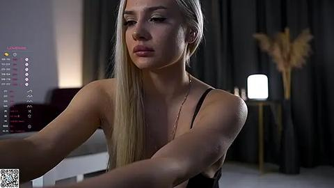 Media: Video of a blonde woman with fair skin, wearing a black tank top, focused on a smartphone screen, in a dimly lit, modern room with dark curtains and a tall plant.