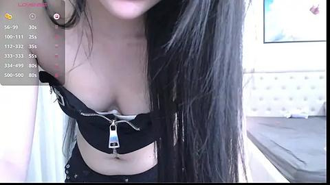 Media: A video shows a topless woman with long black hair, wearing a black bra with a zipper partially undone, revealing her breasts. The background features a bedroom with a white headboard and a bed.