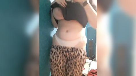 Media: Video of a woman lifting her shirt to reveal her white lace bra and leopard-print pajama pants, standing in a teal room with a blurred background.