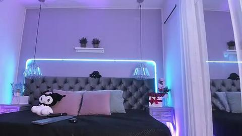 Media: Video of a modern, minimalist bedroom with a plush, gray tufted headboard, two potted plants, and a plush toy on a black bedspread. Purple and blue ambient lighting highlights the room's chic decor.