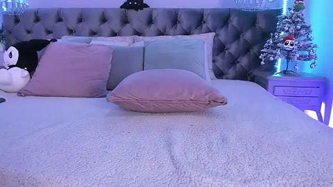 Media: Video of a plush, cushioned bed with a gray, tufted headboard, pink pillow, and plush toys. Nightstand features a Christmas tree with ornaments.