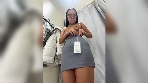 Media: Video of a topless woman with glasses, wearing a gray skirt, holding a white name tag, standing in a dressing room with white curtains and hanging clothes.