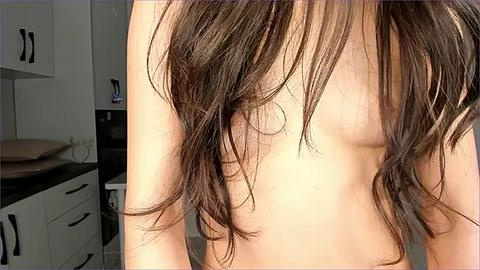 Media: A close-up video of a woman's bare back and shoulder, showing dark brown, wavy hair cascading over her shoulder. The background features a modern kitchen with white cabinets and black handles.