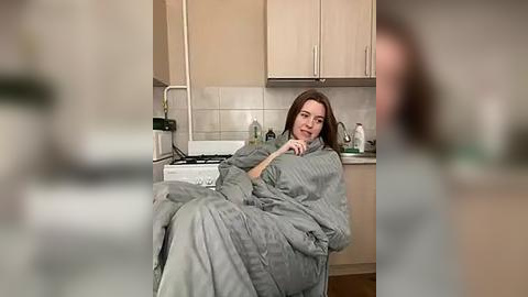 Media: Video of a woman in a gray bathrobe sitting on a kitchen counter, leaning against the wall, with a soft smile, in a modern kitchen with light cabinets and white tiles.