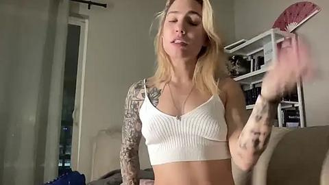 Media: Video of a blonde woman with tattoos on her arm, wearing a white ribbed crop top, smiling, holding a pink feather fan, in a cozy living room with shelves and a window.