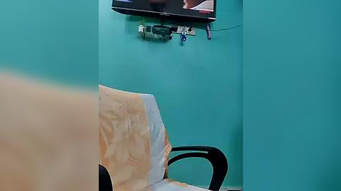 Media: A video shows a teal-painted wall with a mounted TV displaying a person's face. Below the TV, a white bedsheet drapes over a black chair, with a black power strip and cables visible.