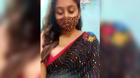 Media: Video of a woman in a black saree with floral embroidery, wearing a face mask, with blurred background.