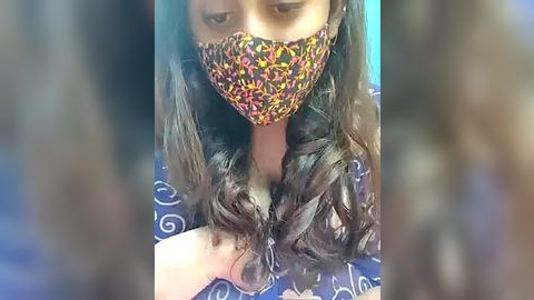 Media: Video of a person with long, wavy, dark brown hair wearing a colorful face mask, holding a section of their hair, against a blue and white patterned background.