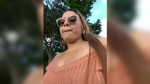 Media: Video of a smiling, light-skinned woman with long, straight brown hair, wearing large, dark sunglasses and a knitted, off-shoulder peach sweater, standing outdoors among greenery.