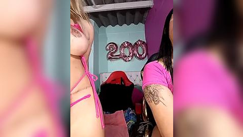 Media: Video of two young women in pink bikinis, one with large breasts, the other with a tattooed arm, standing in a cluttered room with \"2009\" decorations.