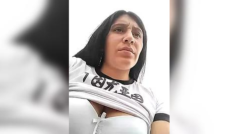 Media: Video of a woman with long black hair and fair skin wearing a white T-shirt, slightly lifted to reveal a white bra. She looks serious.