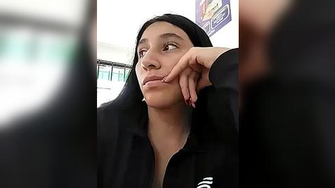 Media: Video of a young woman with medium skin tone and long black hair, wearing a black jacket, resting her head on her hand in contemplation. Background features a blurred office setting with green and white decor.