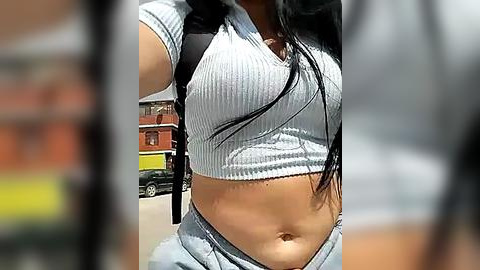 Media: Video of a young woman with medium-dark skin, wearing a tight, ribbed, light gray crop top and light blue jeans, capturing her midsection. Background features blurred urban elements, including a red brick building and a black car.